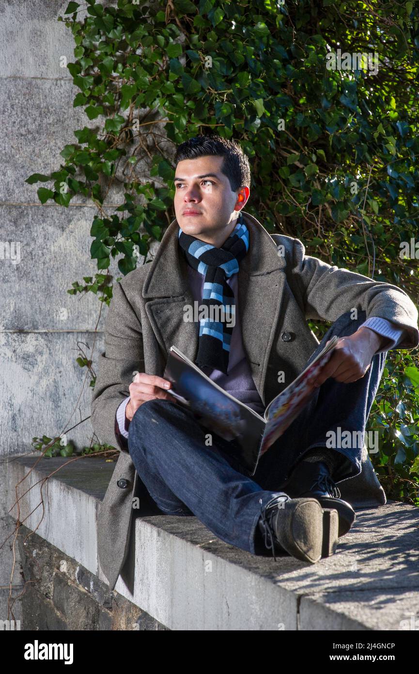 Ein junger Mann, der auf einem großen Rahmen sitzt und vor wilden grünen Pflanzen sitzt, liest ein Magazin und denkt nach. Stockfoto