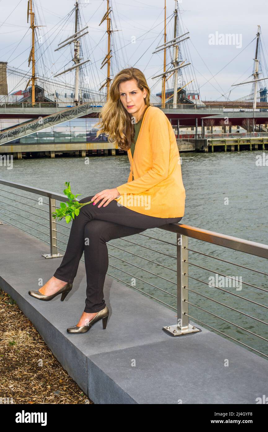 Eine junge, hübsche Frau, die grüne Pflanzen hält, sitzt auf einem Zaun am Fluss und denkt tief nach. Hintergrund ist ein Hafen. Stockfoto