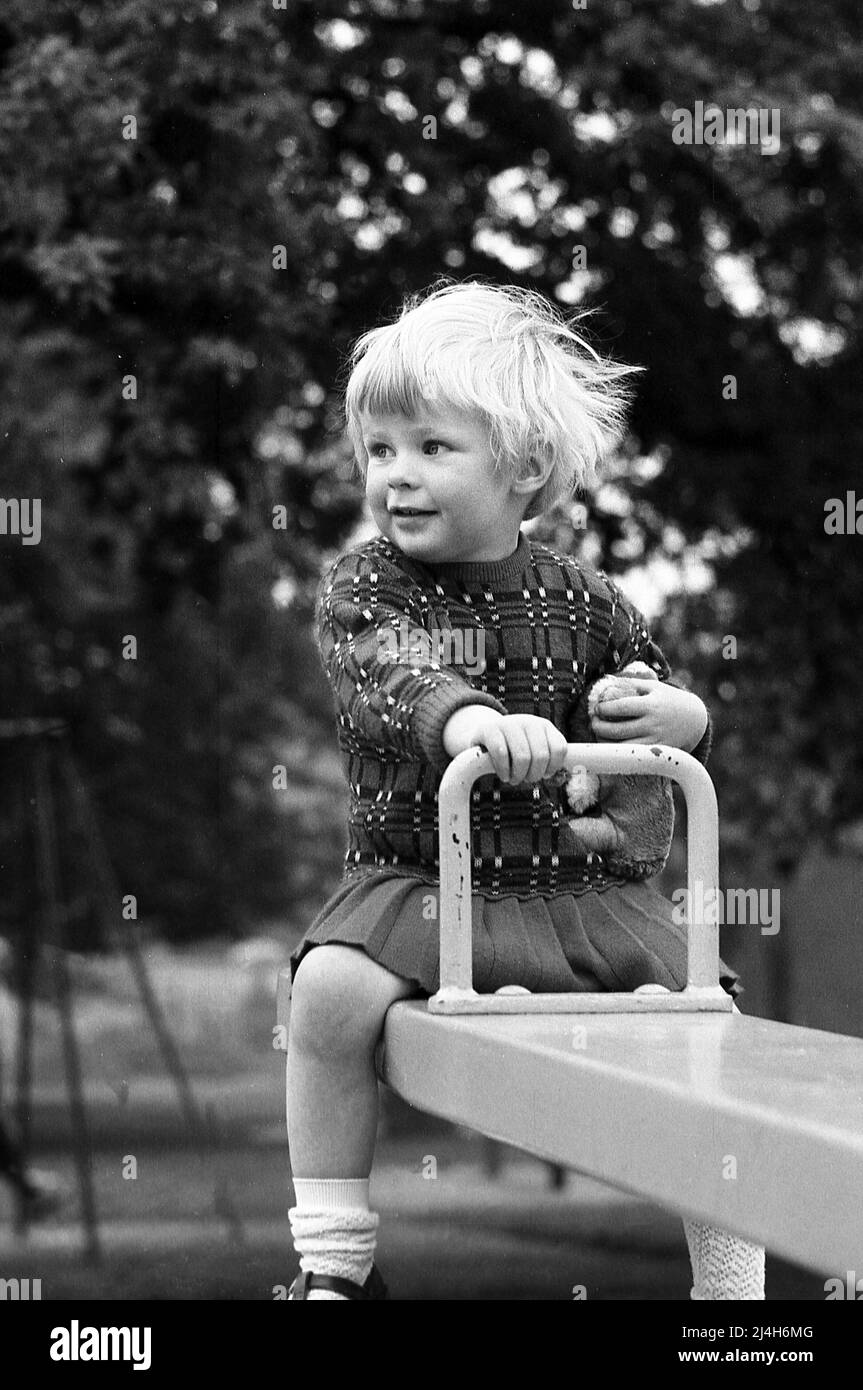 1969, historisch, sitzt ein kleines Mädchen auf einem Spielplatz Wippe, eine Hand hält den Metallgriff, die andere, auf ihr weiches Spielzeug, England, Großbritannien. Stockfoto