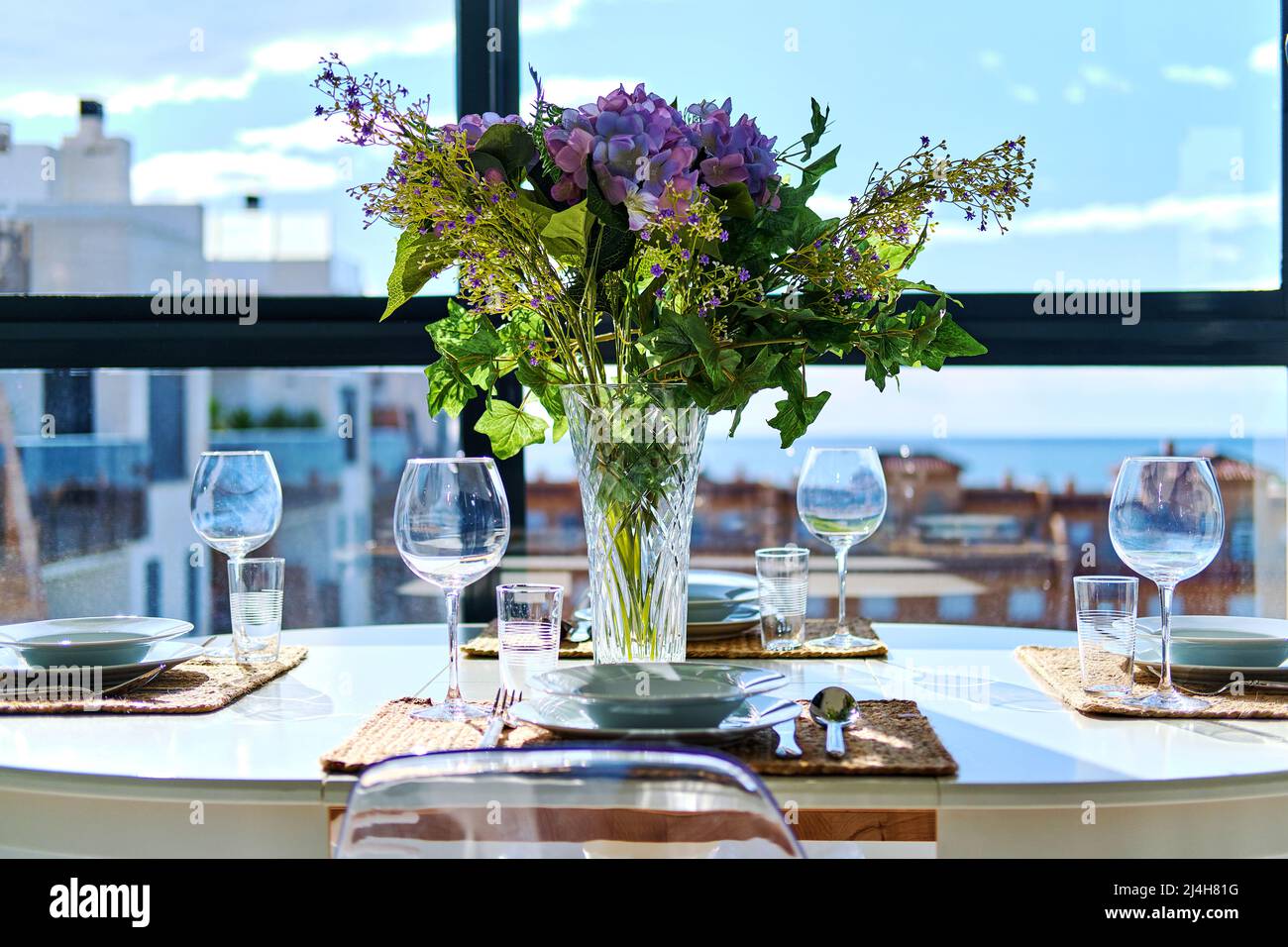 Schöne Tischanordnung mit Blumenstrauß in Vase, serviert Plätze mit Geschirr, Meer und Häuser Dächer Blick durch Fenster Stockfoto