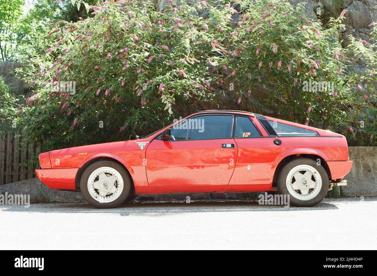 Ein alter roter Sportwagen parkt vor einem grünen Garten. Hintergrund an einem Mittag, helle, warme Sommeraufnahmen, aufgenommen mit einer analogen Filmkamera, die eine sommerliche Atmosphäre verleihen Stockfoto