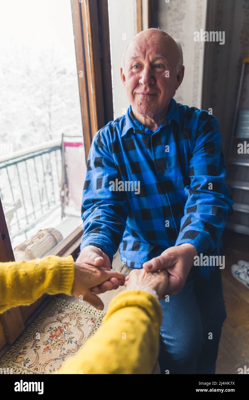 Vertikale Aufnahme eines älteren kaukasischen Rentnermanns, der die Hände seiner Frau am Fenster hielt. Hochwertige Fotos Stockfoto