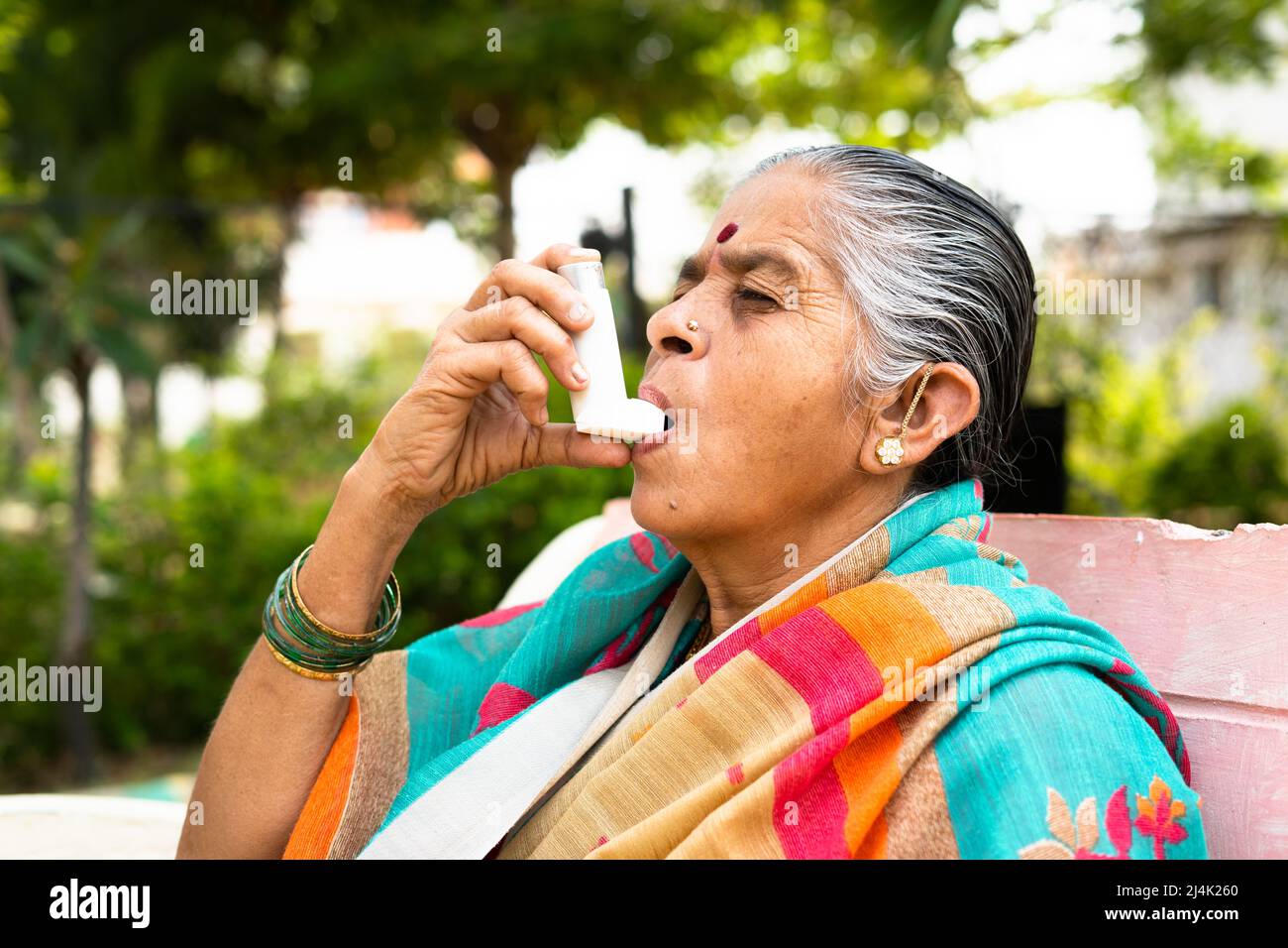 Ältere Frau, die aufgrund einer Allergie Asthma-Inhalator verwendet, während sie im Park sitzt - Konzept, das Auswirkungen von Verschmutzung, Krankheit und Krankheit zeigt Stockfoto