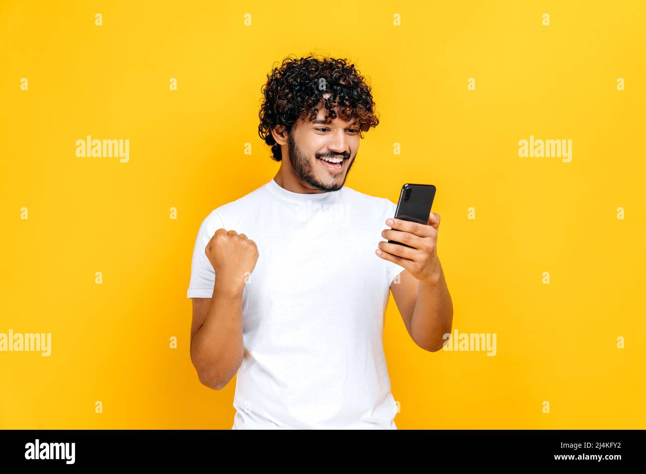 Glücklich fröhlich aufgeregt indischen oder arabischen Kerl hält Smartphone, erhalten unerwartete Nachrichten, gewinnen Lotterie, steht auf isolierten orangefarbenen Hintergrund, fröhlichen Gesichtsausdruck, toothy Lächeln, gestikulieren mit Faust Stockfoto