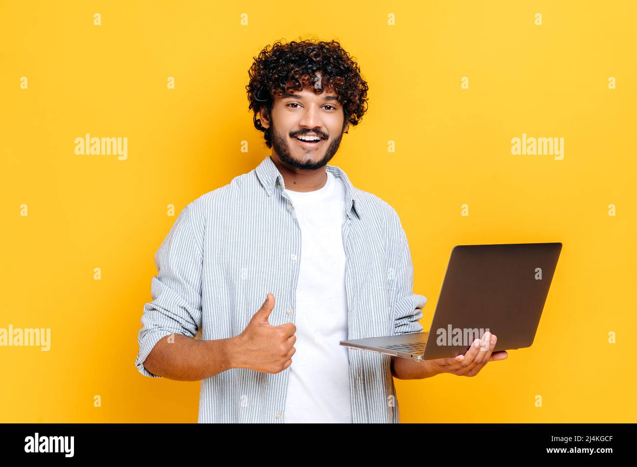 Positiv attraktiver arabischer oder indischer Mann mit lockigem Haar, in legerer Kleidung, mit offenem Laptop in der Hand, blickt auf die Kamera, zeigt Daumen nach oben, steht auf isoliertem orangefarbenem Hintergrund Stockfoto