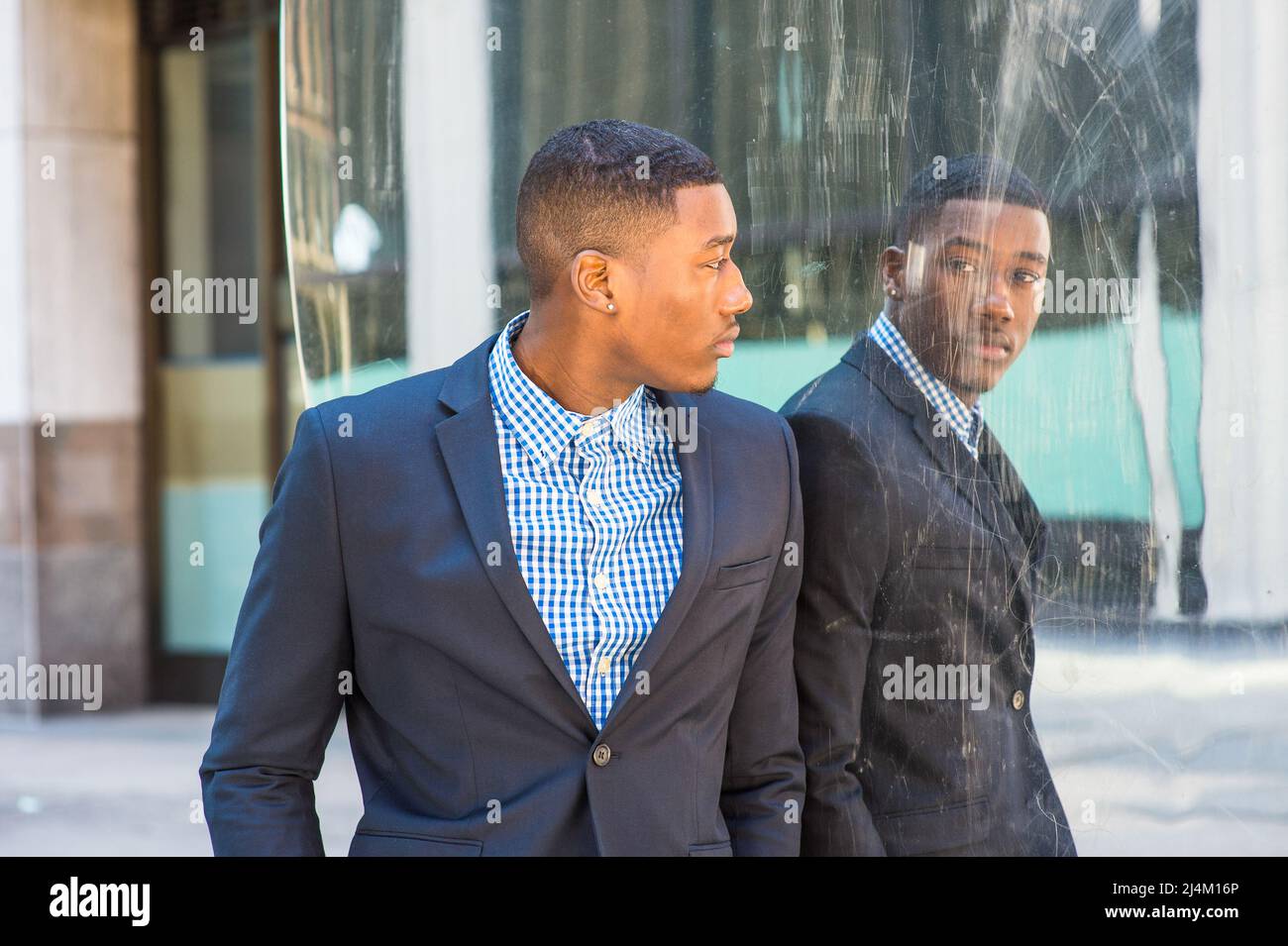 Junger afroamerikanischer Geschäftsmann in blauer Jacke, gemustertem Hemd, beim Spiegel stehend, sich umdrehend, Spiegelbild anblickend, Denken. Konzept Stockfoto
