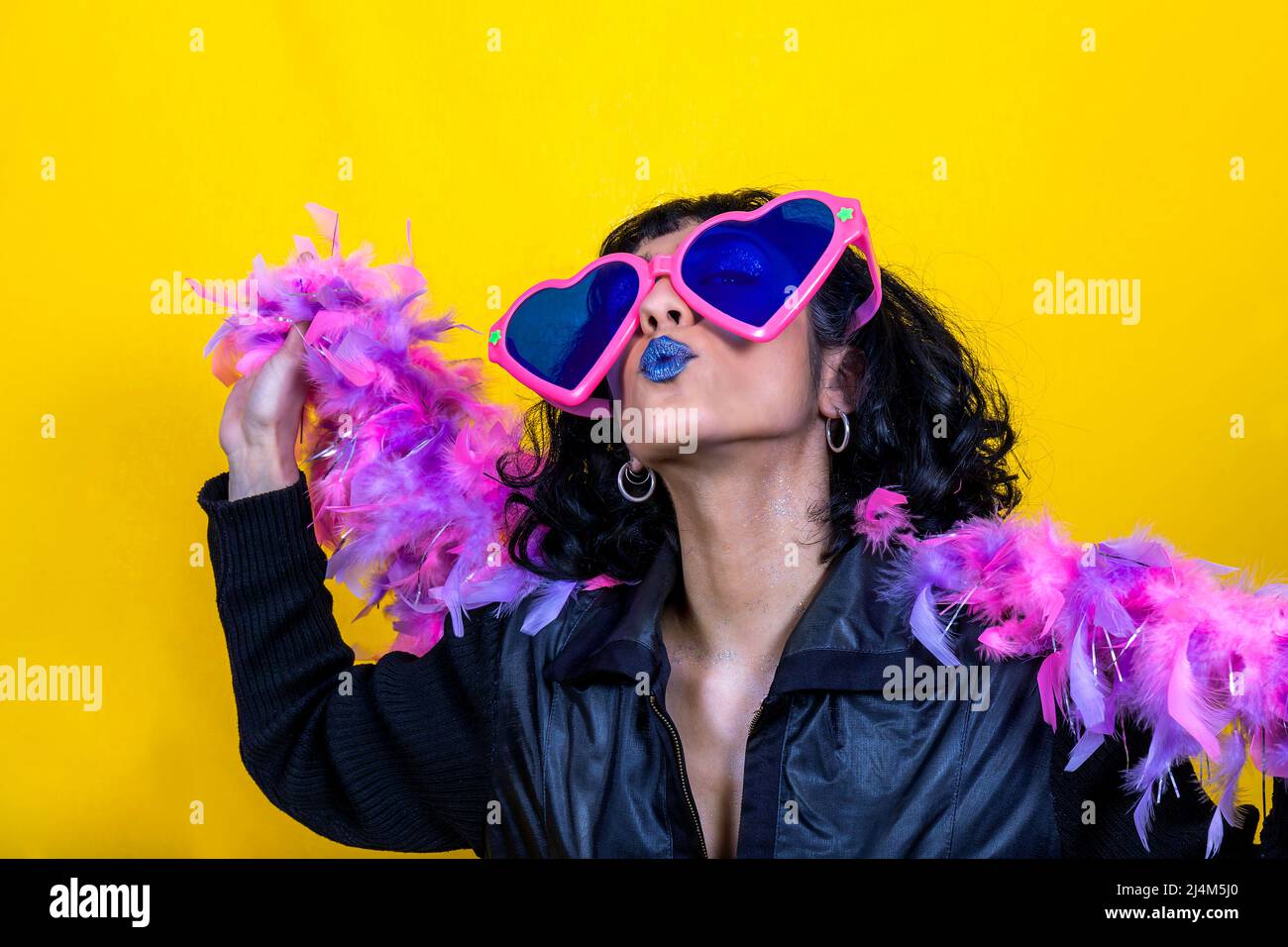 Die junge Frau trägt eine große herzförmige Brille und einen Schal mit rosa und violetten Federn. Schöne Frau schickt einen Kuss. Party-Konzept. Stockfoto