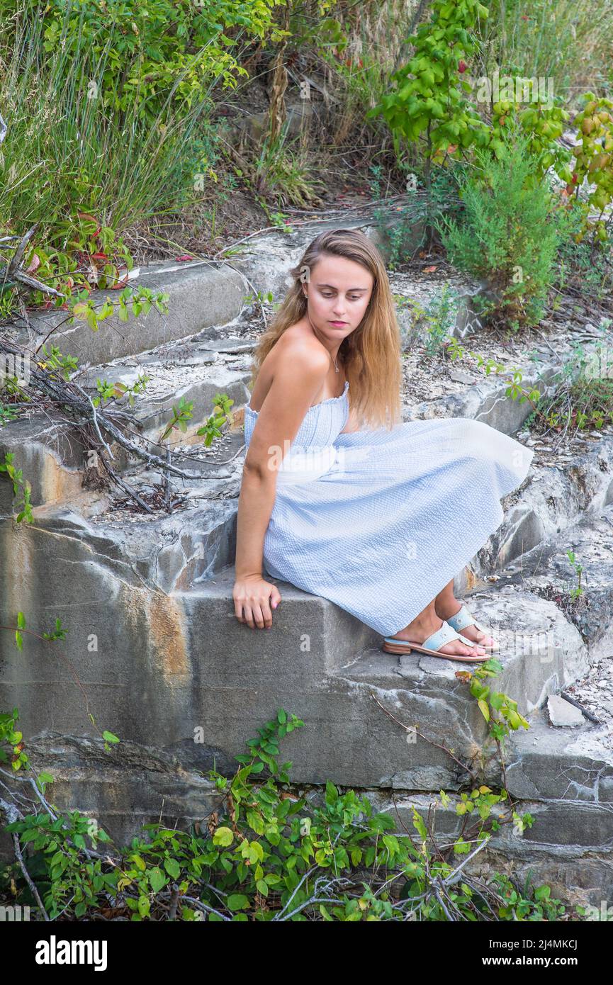 Ein hübsches blondes Mädchen, das sich in einem weißen blauen Kleid, weißem Bund und Sandalen kleidet, sitzt auf einem wilden, isolierten Feld, um tief zu denken. Stockfoto