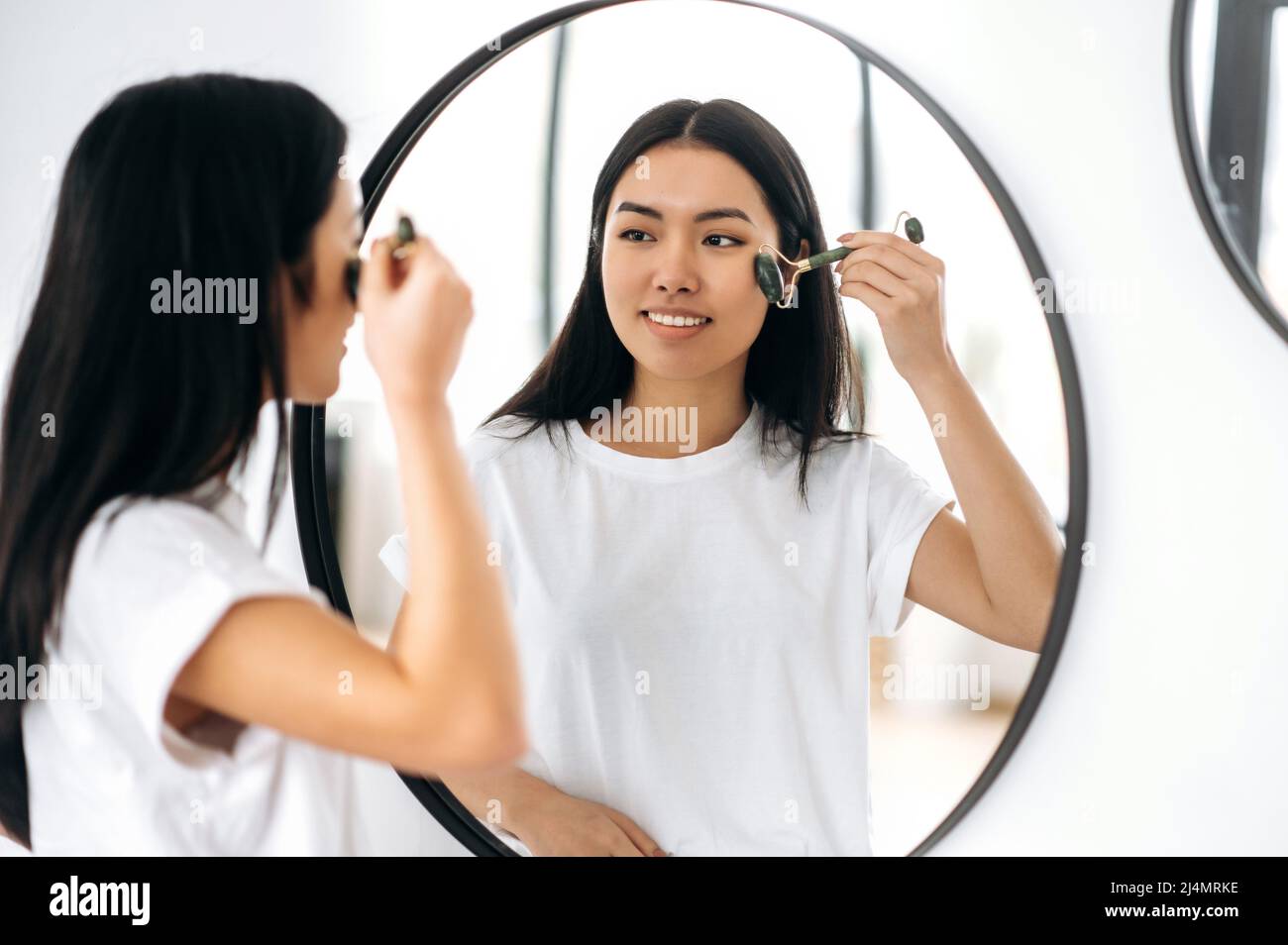 Happy Asian Brunette hübsches Mädchen im T-Shirt, steht zu Hause vor einem Spiegel, macht ihre morgendliche Schönheitsroutine, kümmert sich um ihre Gesichtshaut, verwendet eine Gesichtssteinrolle, beugt Falten vor, lächelt Stockfoto