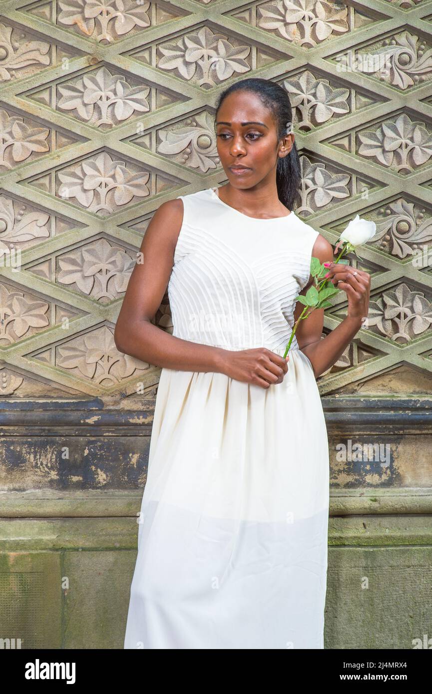 Ein junges, hübsches schwarzes Mädchen, das sich in Weiß kleidet und eine weiße Rose hält, steht an einer Wand im uralten Stil und denkt tief nach. Stockfoto