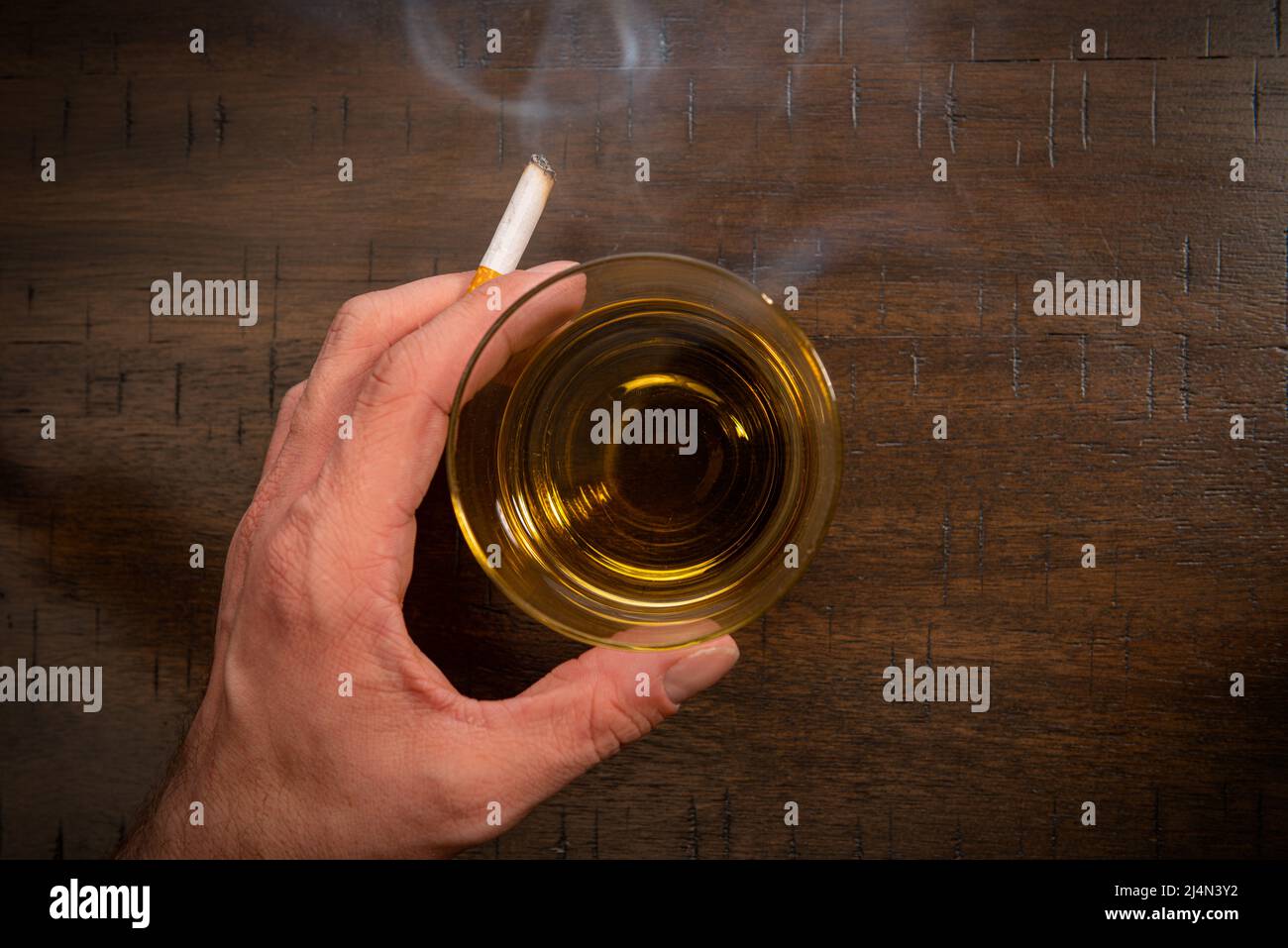 Eine männliche kaukasische Hand hält eine rauchende Zigarette in der Nähe eines Glases Whisky. Stockfoto