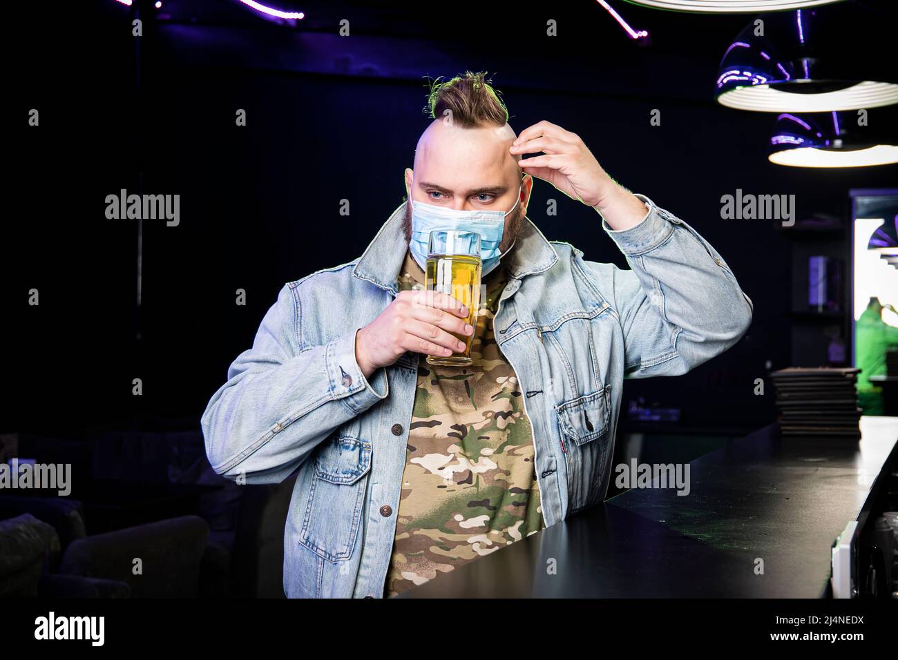 Ein Punk-Mann mit einem Mohawk, der eine Schutzmaske auf seinem Gesicht  trägt, trinkt im Dunkeln Bier aus einem Glaskrug. Quarantäne in der Leiste.  Alkoholverbot Stockfotografie - Alamy
