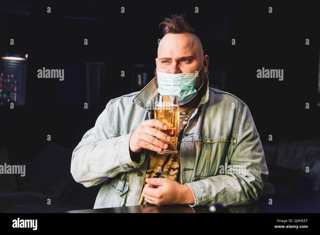 Ein Punk-Mann mit einem Mohawk, der eine Schutzmaske auf seinem Gesicht trägt, trinkt im Dunkeln Bier aus einem Glaskrug. Quarantäne in der Leiste. Alkoholverbot Stockfoto