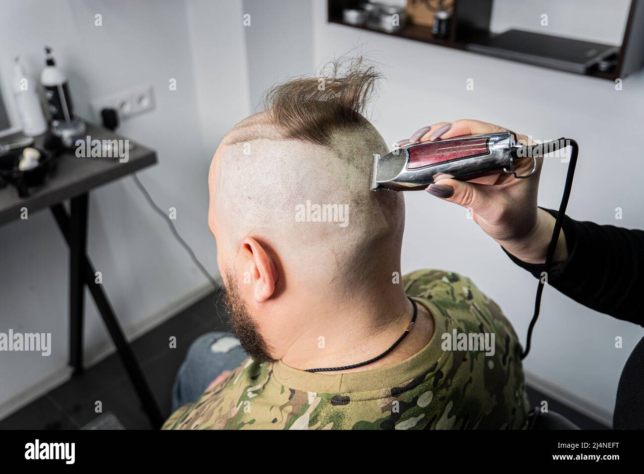 Ein junger Mann in Militäruniform rasiert sich zum Militärdienst den Kopf kahl. Ein Mann mit Bart bekommt einen Haarschnitt in einem Friseurladen. Stockfoto