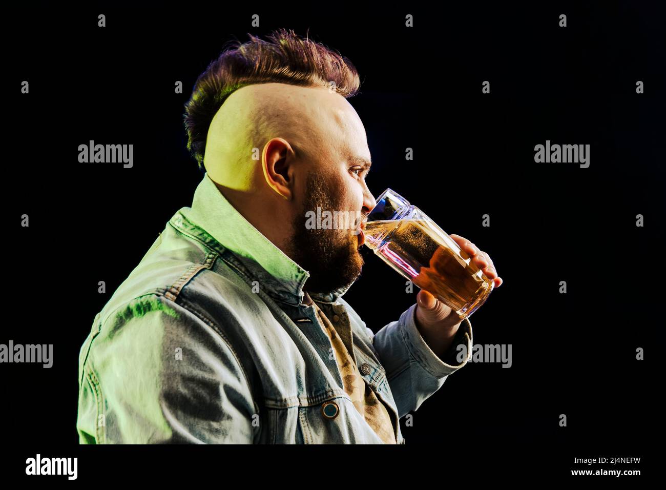 Ein Punk-Mann mit einem Mohawk trinkt im Dunkeln Bier aus einem Glaskrug. Das Konzept der Alkoholabhängigkeit. Stockfoto