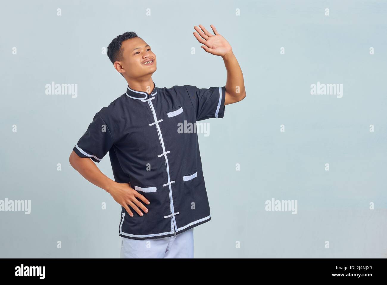 Porträt eines jungen, schmunzelten asiatischen Mannes, der Taekwondo Kimono trägt und eine winkende Geste mit isolierten Handflächen auf grauem Hintergrund macht Stockfoto