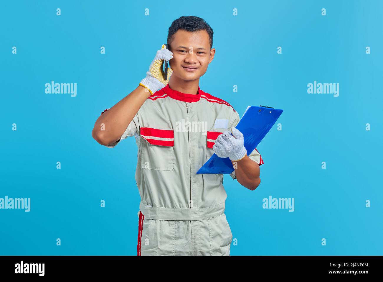 Lächelnder junger asiatischer Mechaniker, der auf dem Handy spricht und Clipboard auf blauem Hintergrund isoliert hält Stockfoto