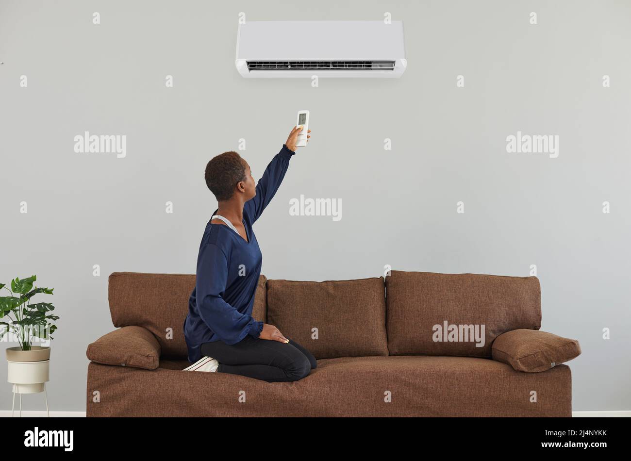 Junge afroamerikanische Frau, die zu Hause auf der Couch sitzt, schaltet die Klimaanlage an der Wand Stockfoto