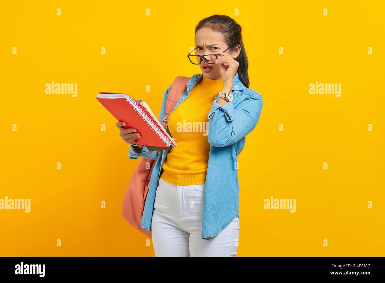 Nachdenklich schöne junge asiatische Frau Student in Denim-Kleidung mit Rucksack halten Notebook und Blick auf Kamera isoliert auf gelbem Hintergrund. Educa Stockfoto