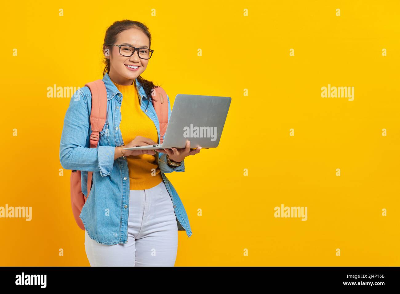 Porträt einer fröhlichen jungen asiatischen Studentin in legerer Kleidung mit Rucksack mit Laptop und Blick auf die Kamera isoliert auf gelbem Hintergrund. Educa Stockfoto