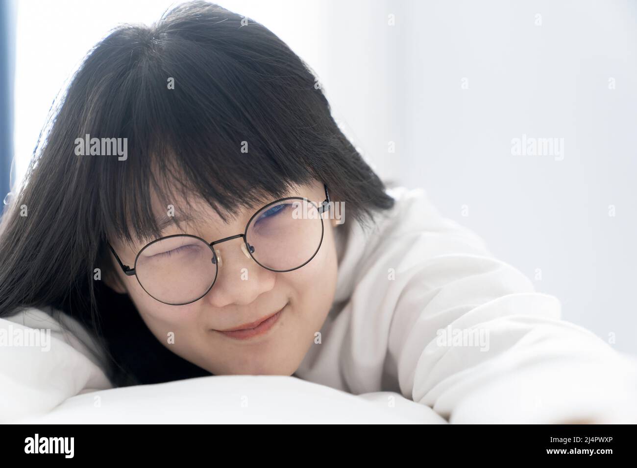 Winked Gläser süß, niedlich asiatische Mädchen legt sich auf das Bett mit glücklich und Komfort Gefühl vor dem Fenster Licht am Morgen. Stockfoto