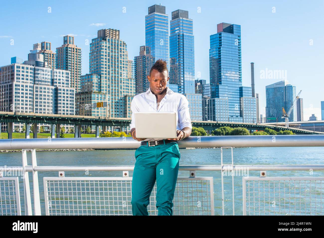 In einem weißen Hemd und einer grünen Hose kleidet, steht ein junger schwarzer Kerl mit Mohawk-Haaren vor hohen Gebäuden am Wasser und arbeitet an einem c Stockfoto