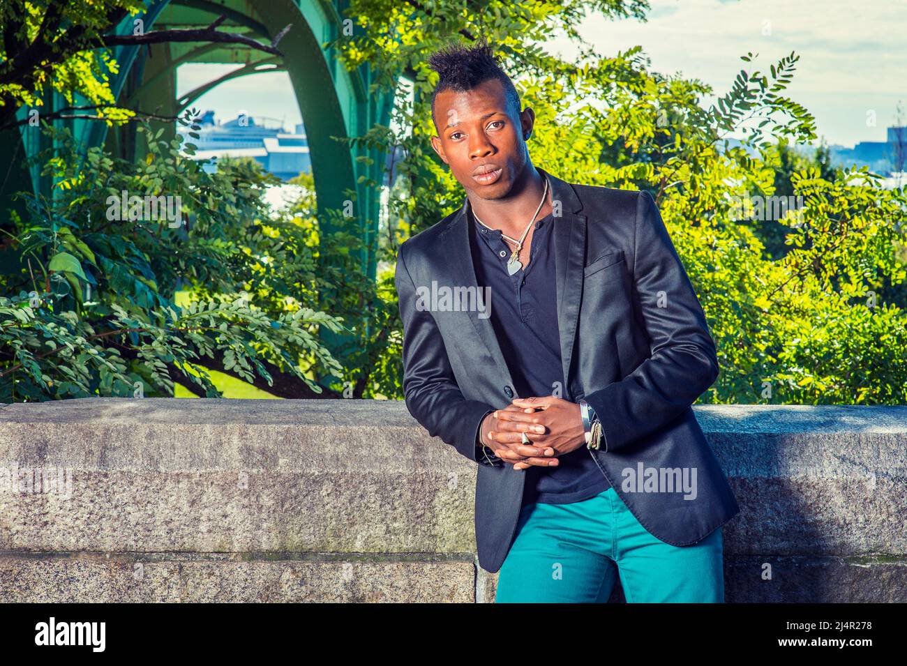 Mann, Der Sich Draußen Entspannt. In einem schwarzen Blazer, einem schwarzen Unterhemd, einer grünen Hose, steht ein junger schwarzer Kerl mit Mohawk-Haaren an einem felsigen Zaun, RE Stockfoto