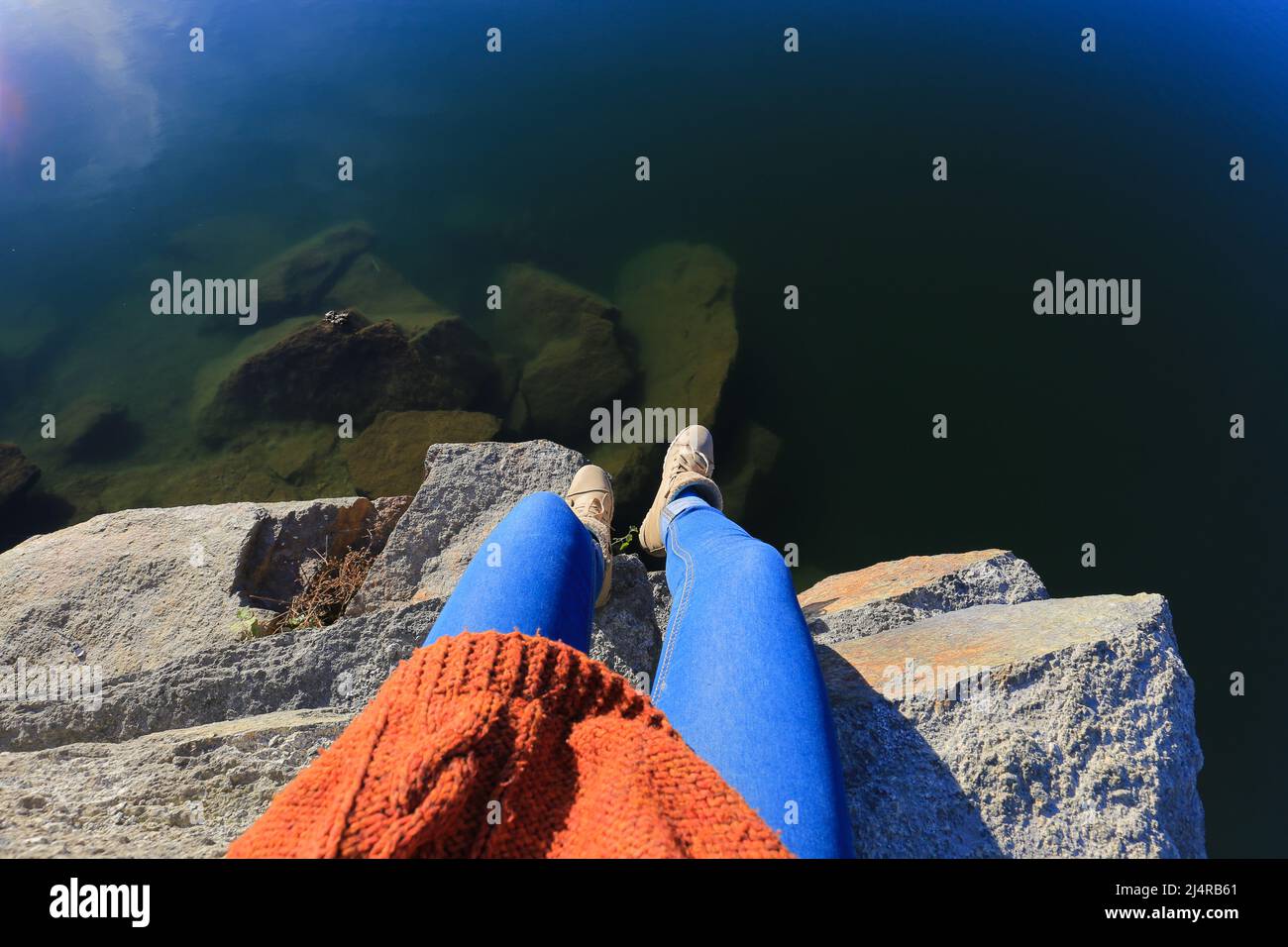 Aus der ersten Person kann man die Beine einer Frau sehen, die auf einer Klippe sitzt und einen atemberaubenden Blick auf den See hat Stockfoto