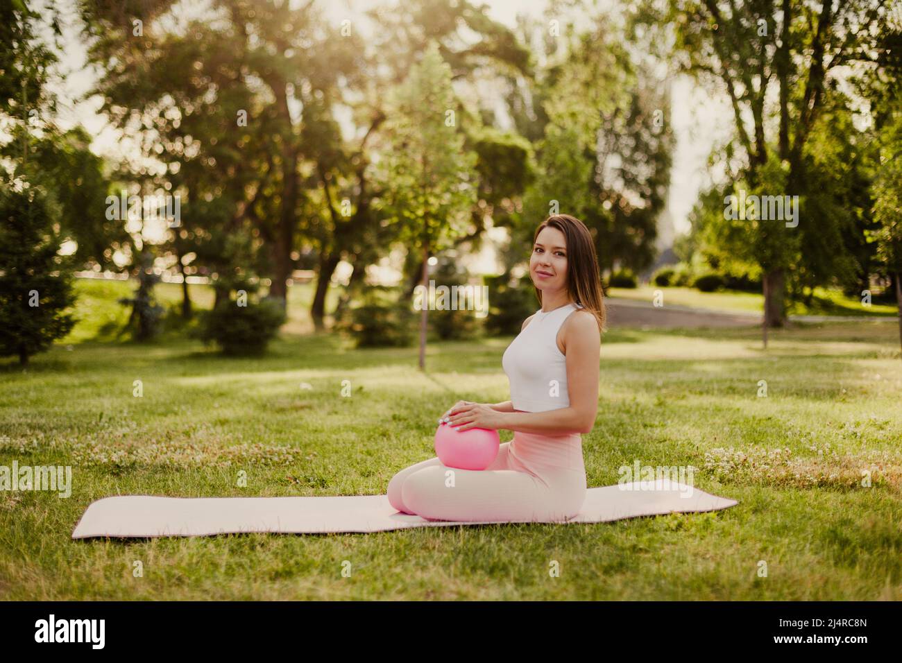 Charmante fit junge Frau sitzt auf den Knien auf Sportmatte mit Gummi kleinen Pilates Ball im Park Stockfoto