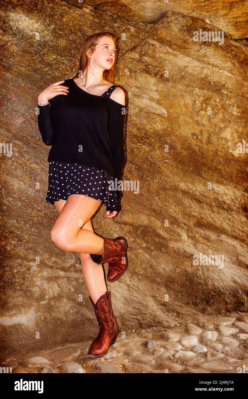 Das amerikanische Teenager-Mädchen trägt einen schwarzen Strickpullover, eine Schulter, einen gemusterten Rock und braune Stiefel und steht im Central Park, New York, gegen die Felswand Stockfoto