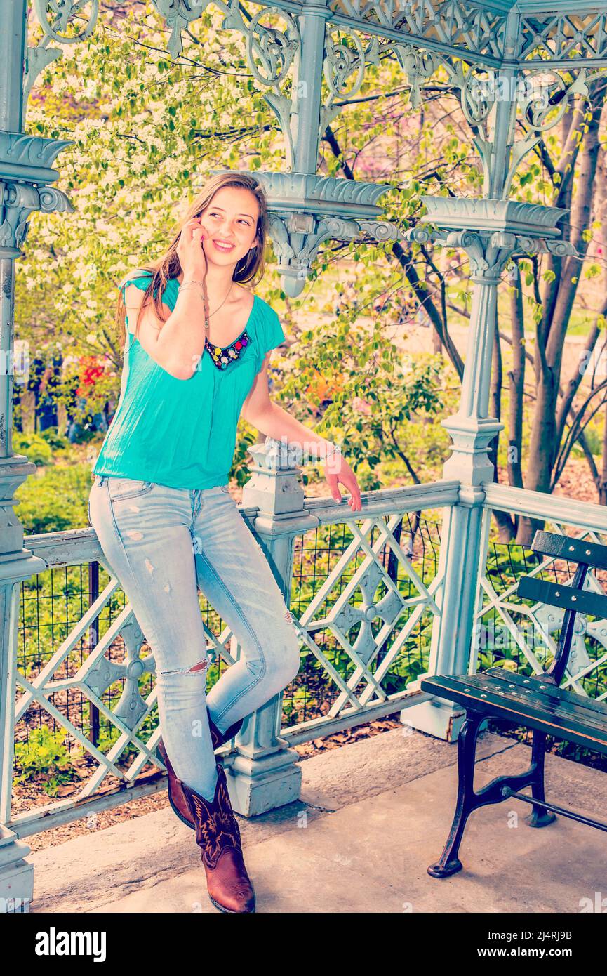 American Teenage girl trägt blaues ärmelloses Top, modische Jeans, braune Stiefel, steht im Pavillon im Central Park, New York, im Frühling Tag, Stockfoto