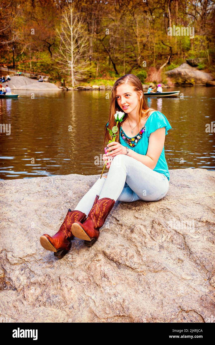Das amerikanische Teenager-Mädchen trägt ein blaues ärmelloses Oberteil, Jeans, braune Stiefel, sitzt auf Felsen am See im Central Park, New York, hält weiße Blume, riecht Stockfoto