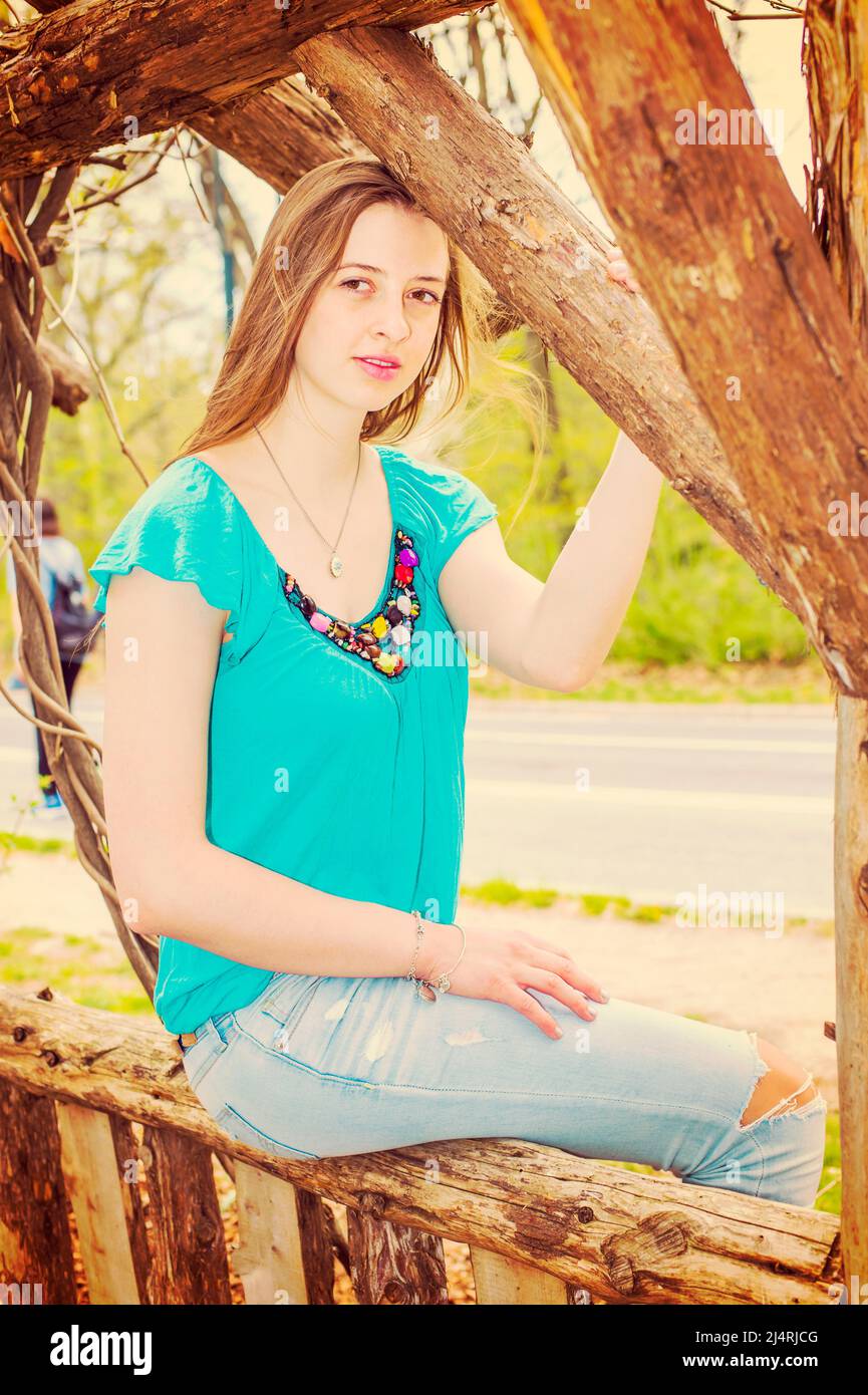 Das amerikanische Teenager-Mädchen trägt ein blaues ärmelloses Oberteil, modische Jeans und sitzt auf einem Holzzaun im Central Park, New York, und blickt auf Sie charmant. Zoll Stockfoto