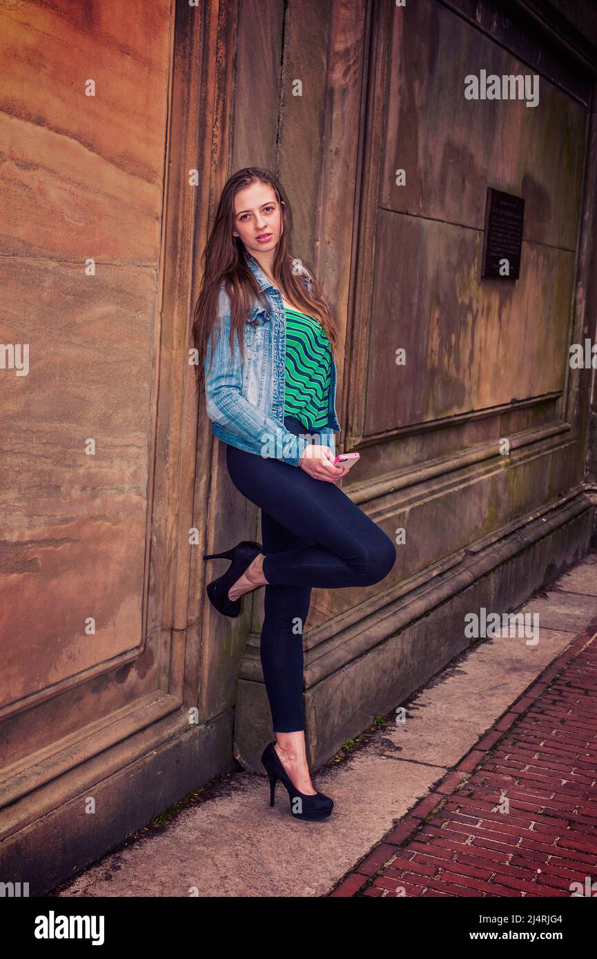 Amerikanische Teenager-Mädchen tragen gemustertes Unterhemd, Denim-Jacke, Legginghose, High heels, Biegebein, Steht an der Vintage Wall in New York, holdin Stockfoto