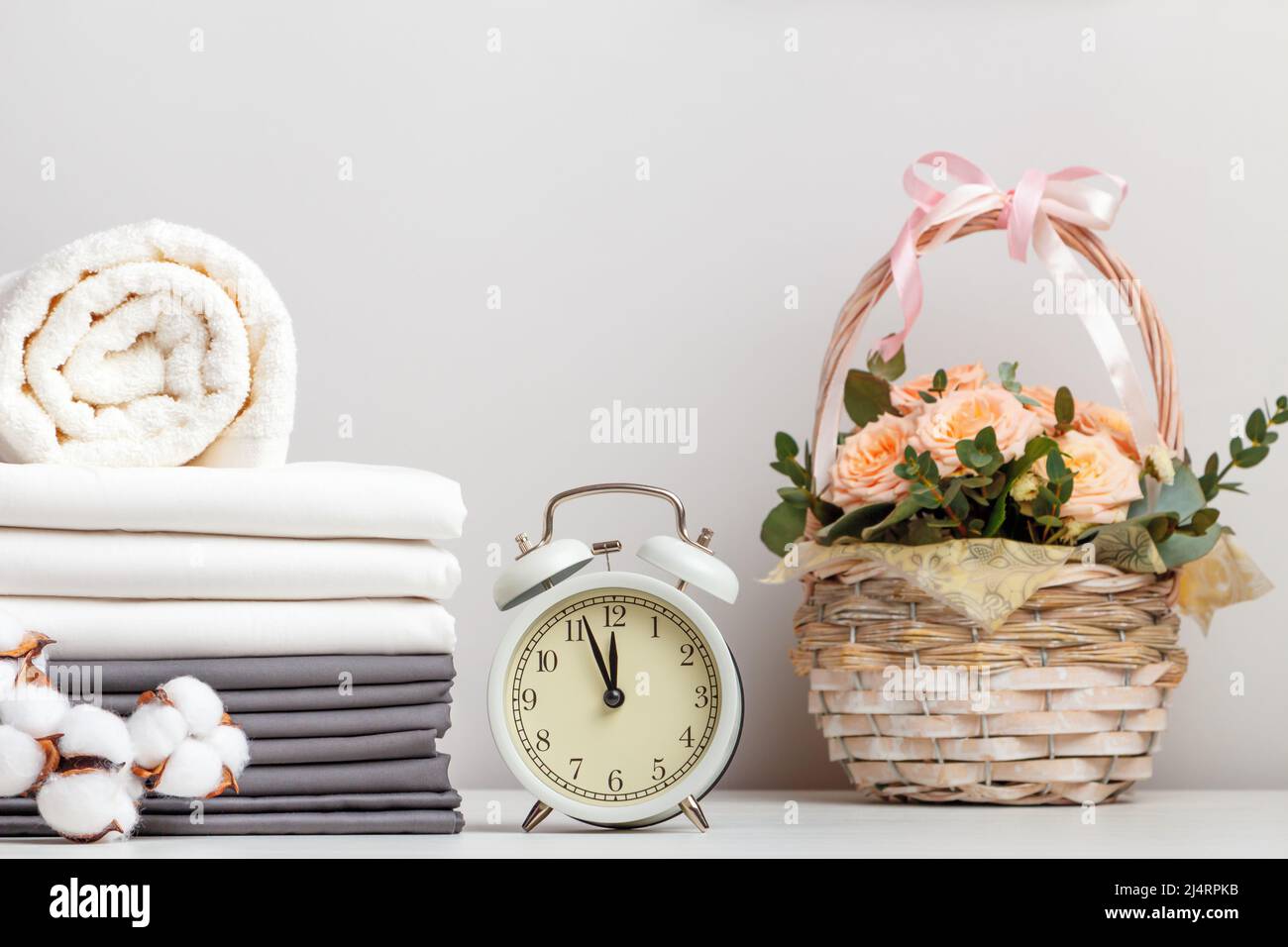 Ein Stapel grauer und weißer Bettwäsche, Laken und ein Frottee-Handtuch. Wecker und ein Blumenkorb auf dem Tisch. Stockfoto