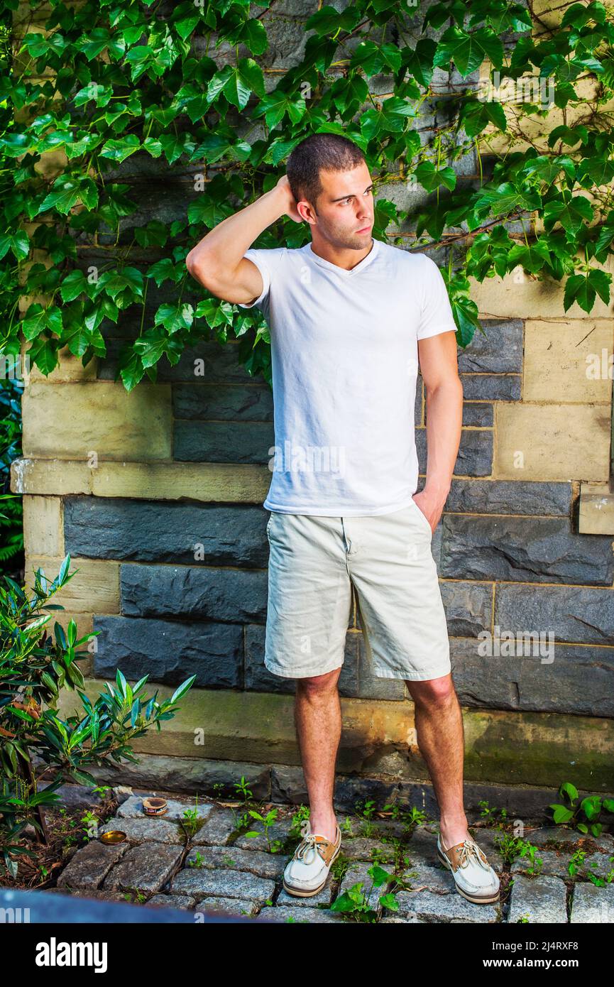 Junger Mann, der denkt. In einem weißen T-Shirt mit V-Ausschnitt, hellgelben Shorts, Lederschuhen, einer Hand, die am Hinterkopf kratzt, steht ein junger Mann b Stockfoto