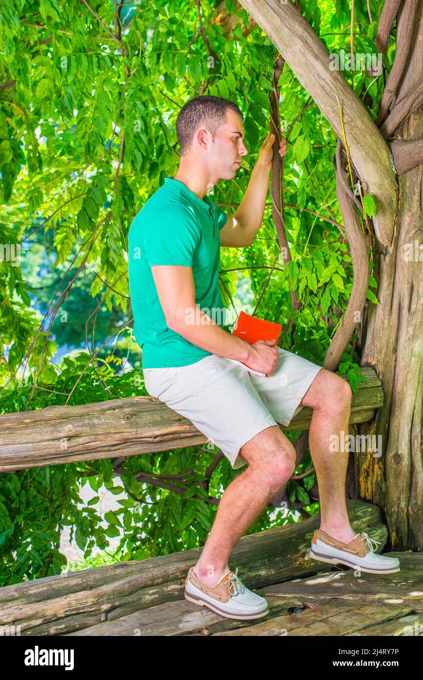 Junger amerikanischer Mann, der nach dem Lesen denkt, trägt grünes Kurzarm-Henley-Hemd, hellgelbe Shorts, Lederschuhe, hält rotes Buch, Sitti Stockfoto