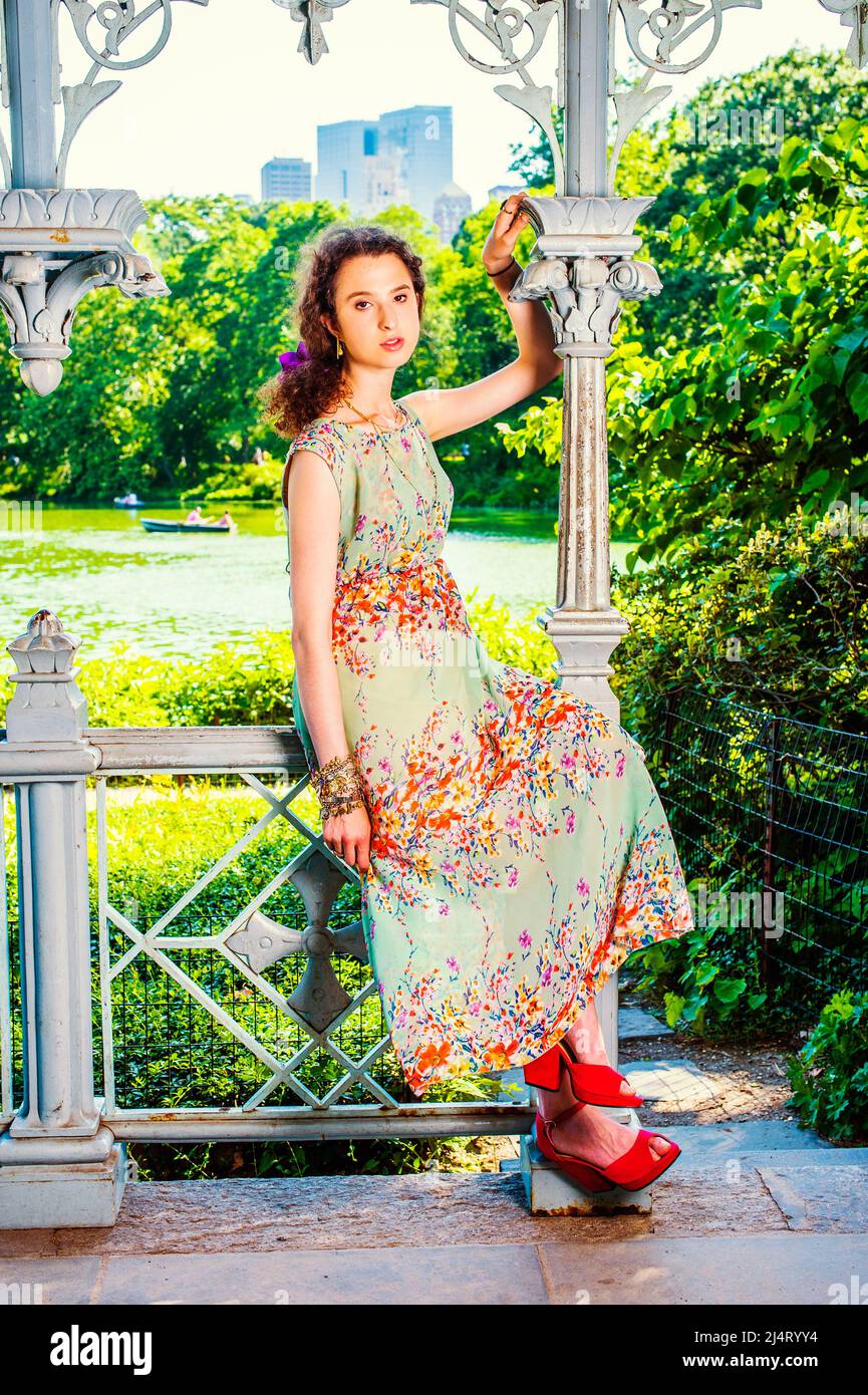 Lady beim Entspannen im Park. Ein hübsches Mädchen im Teenageralter sitzt auf einem Geländer in einem Pavillon und wartet in ärmellosem, langem Kleid, roten Sandalen-Schuhen auf Stockfoto