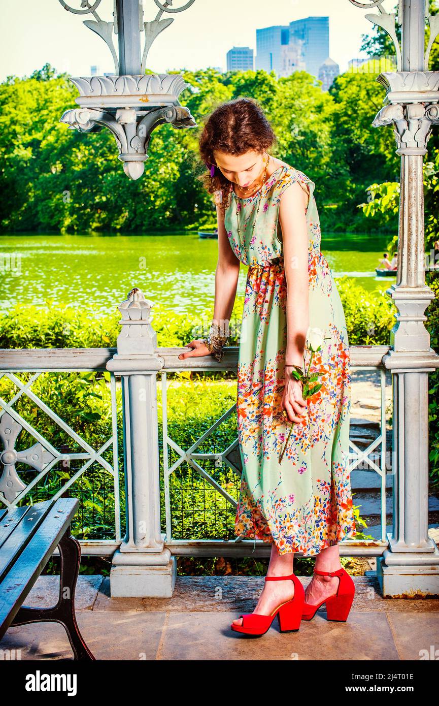 Sommermode. In ärmellosen, langen Kleidern, roten Sandalen-Schuhen kleidet, steht ein Teenager-Mädchen in einem Garten, eine Hand mit einer weißen Rose, loweri Stockfoto