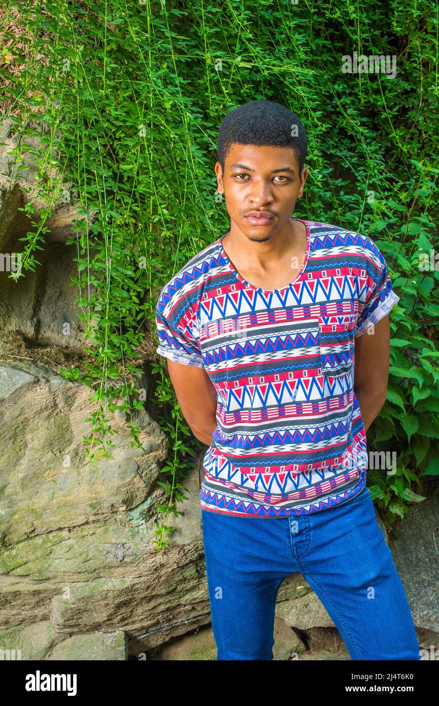 Afroamerikanischer Mann Casual Fashion in New York. Mit bunt gemusterten, kurzen Ärmeln, kragenlosem Hemd, blauen Jeans und einem jungen Mann, der am Fels steht Stockfoto