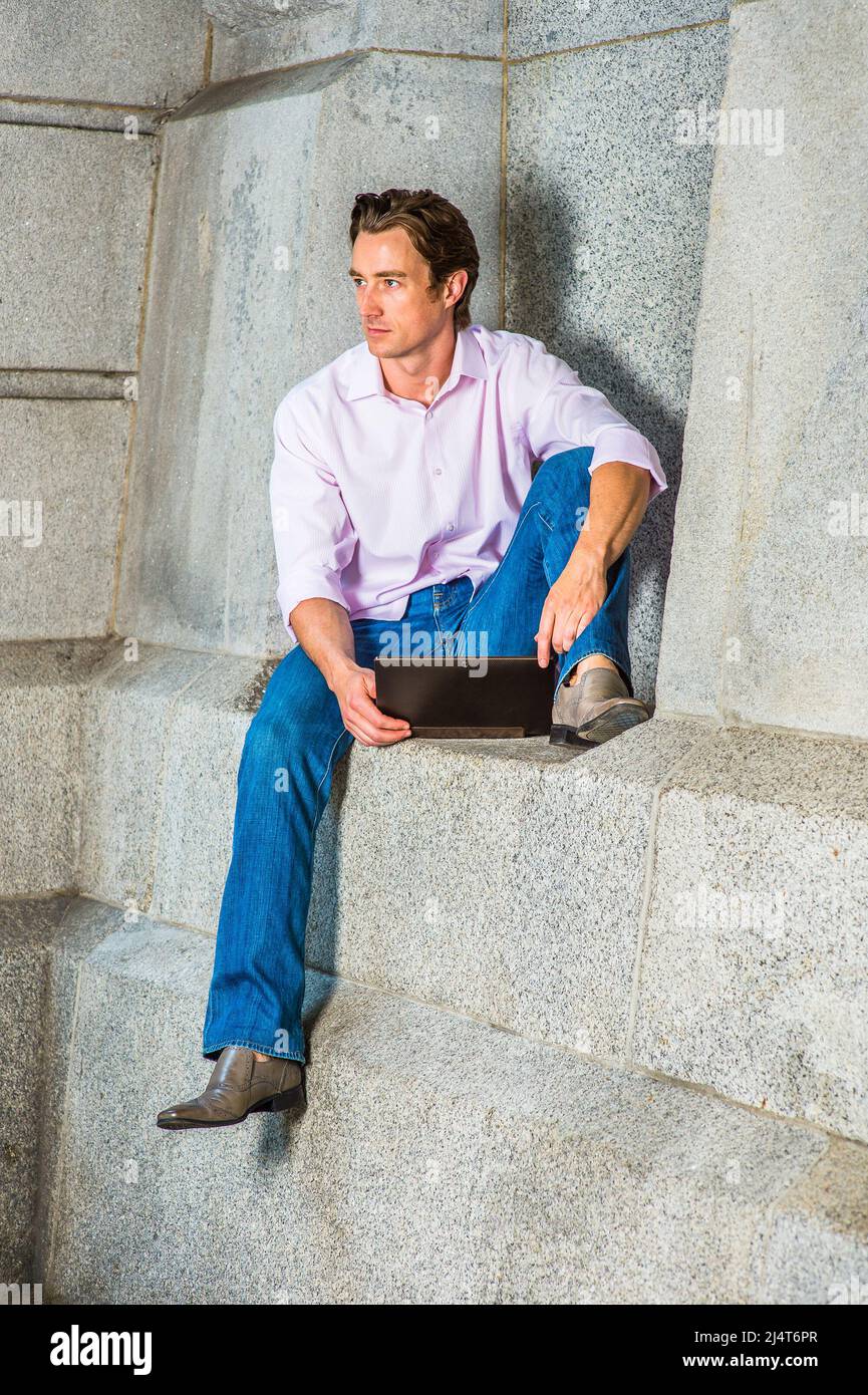 Mann, Der Draußen Arbeitet. Ein junger Geschäftsmann sitzt in einem hellrosa, langärmeligen Hemd, blauen Jeans und Lederschuhen lässig gegen eine Betonwa Stockfoto