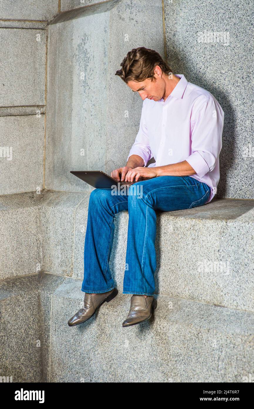 Mann, Der Draußen Arbeitet. Ein junger Geschäftsmann sitzt in einem hellrosa, langärmeligen Hemd, blauen Jeans und Lederschuhen lässig gegen eine Betonwa Stockfoto