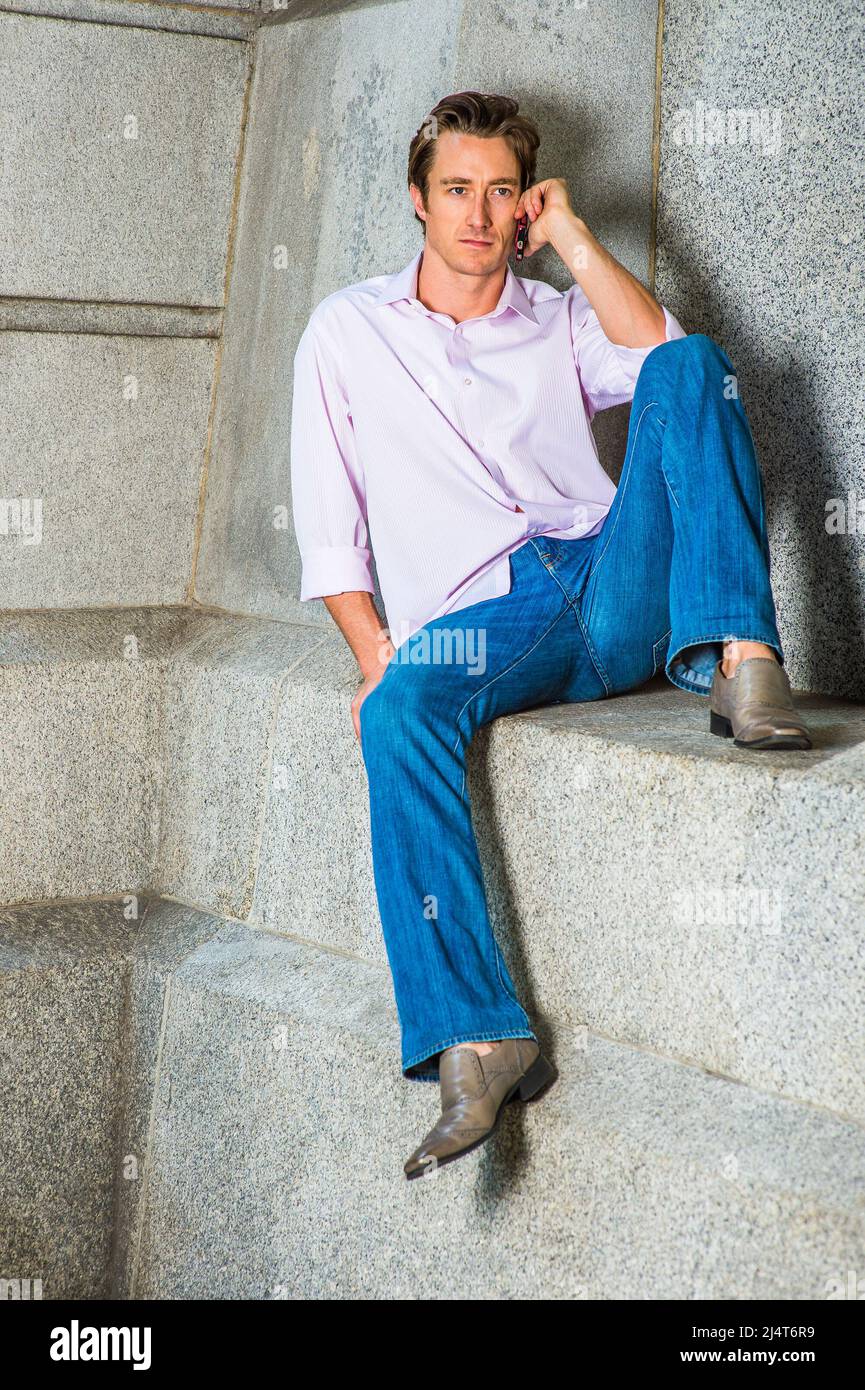 Mann, der am Telefon anruft. In einem hellrosa Langarmshirt, einer blauen Jeans und Lederschuhen sitzt ein junger Geschäftsmann lässig gegen einen Betonklotz Stockfoto