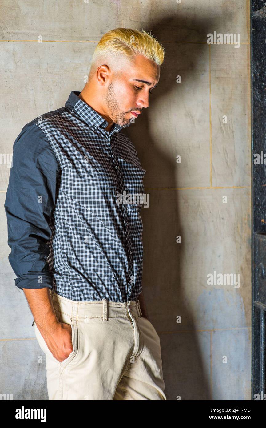 In einem schwarz gemusterten Hemd, einer gelben Hose, einem jungen Mann mit Bart, gelbes Haar lehnt sich vor einem Metalltor an die Wand, Hände in Taschen, Stockfoto