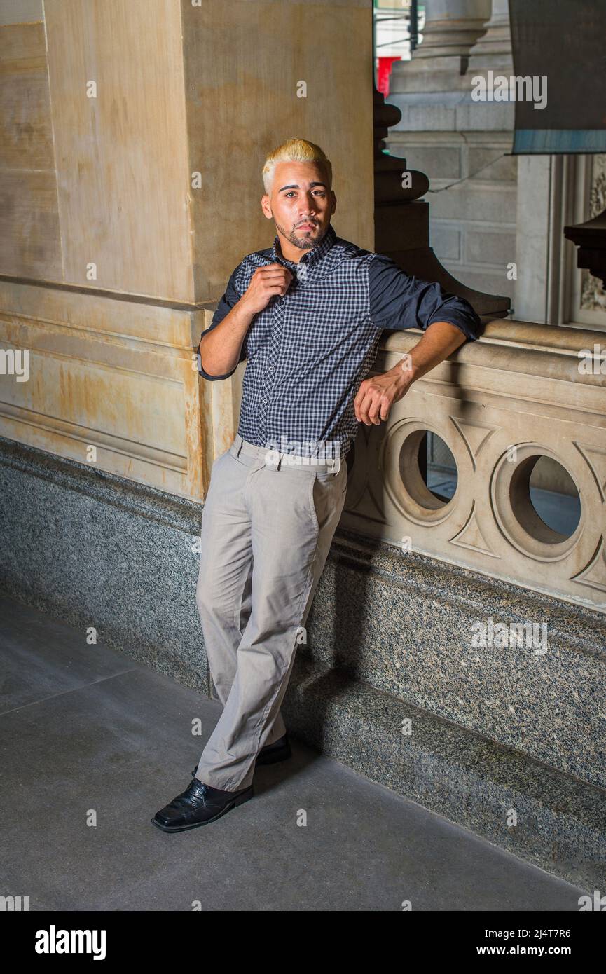Porträt eines jungen Arbeiters. In einem schwarz gemusterten Hemd, einer grauen Hose, Lederschuhen, steht ein junger Mann mit Bart, gelben Haaren an einem Geländer Stockfoto