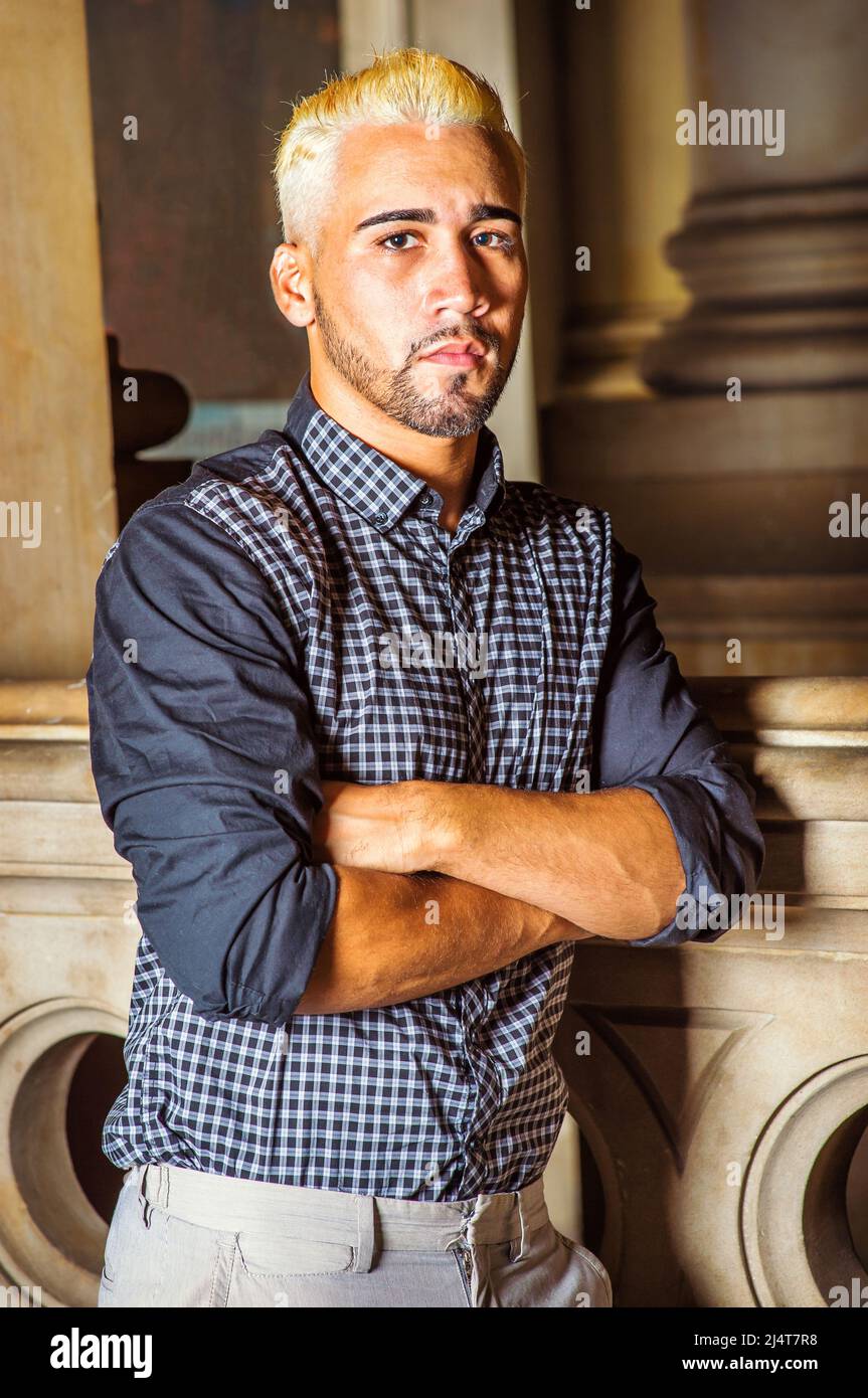 Porträt eines jungen Arbeiters. In einem schwarz gemusterten Hemd, einer grauen Hose, überkreuzenden Armen steht ein junger Mann mit Bart, gelben Haaren, an einem Geländer Stockfoto