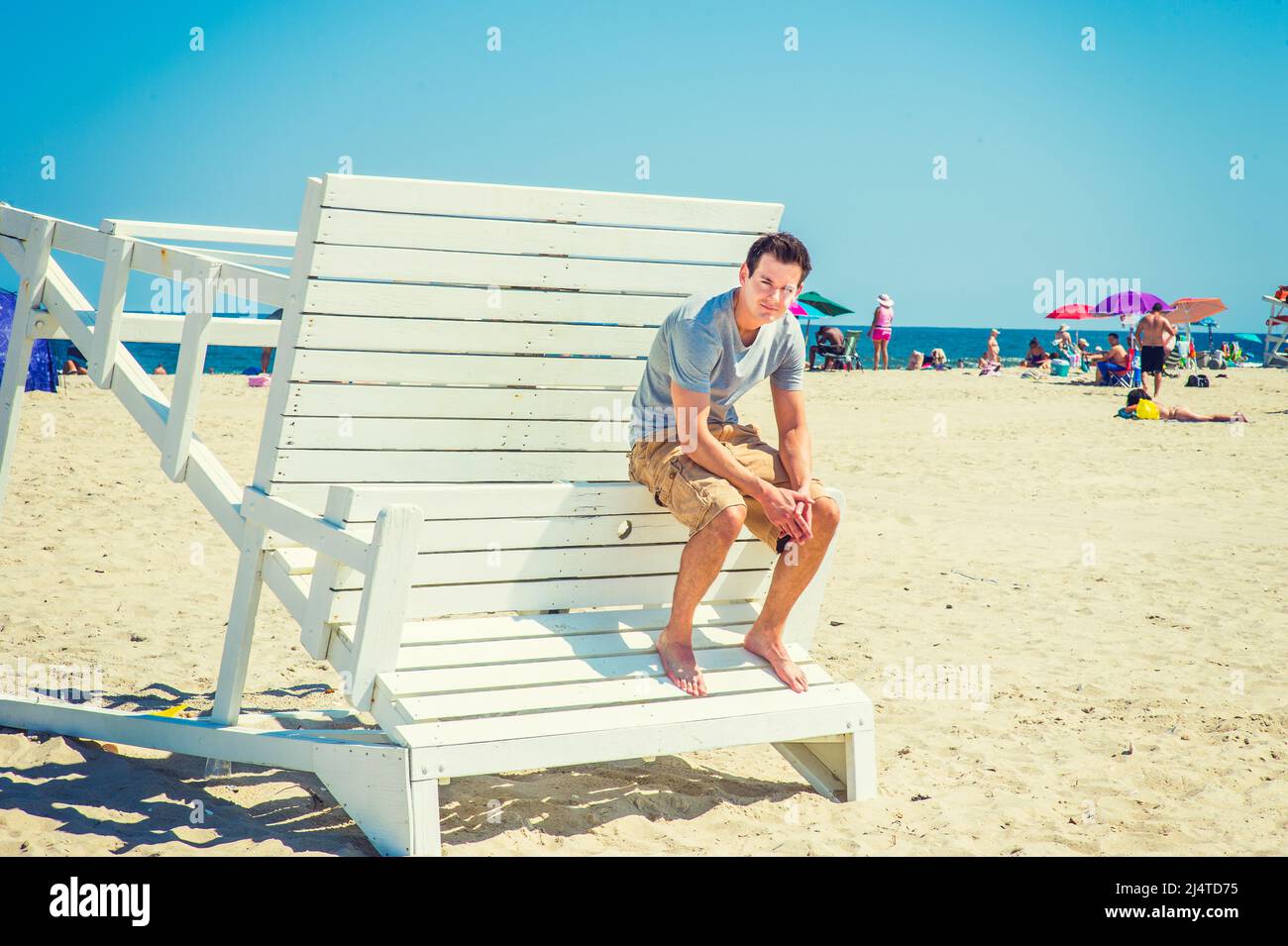 Menschen am Strand. Trägt ein graues T-Shirt, dunkelgelbe Shorts, barfuß, sich nach hinten beugend, Hände haltend, Ein junger, gutaussehender Mann sitzt auf einem Wald Stockfoto