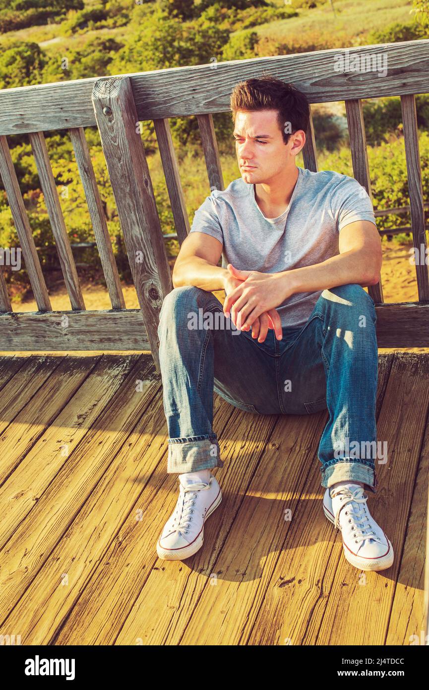 Mann, Der Nach Draußen Denkt. In einem grauen T-Shirt, Jeans und weißen Sneakers sitzt ein junger, hübscher Kerl auf dem Holzboden, zurück gegen den Zaun in einem remo Stockfoto