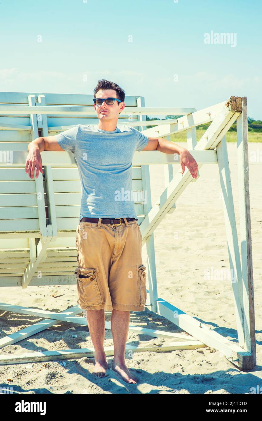 Mann Sommer Casual Fashion. In einem grauen T-Shirt, einer lässigen kurzen Hose, einer Sonnenbrille und auf einem Holzstock ruhenden Armen steht ein junger, gutaussehender Kerl zur Verfügung Stockfoto
