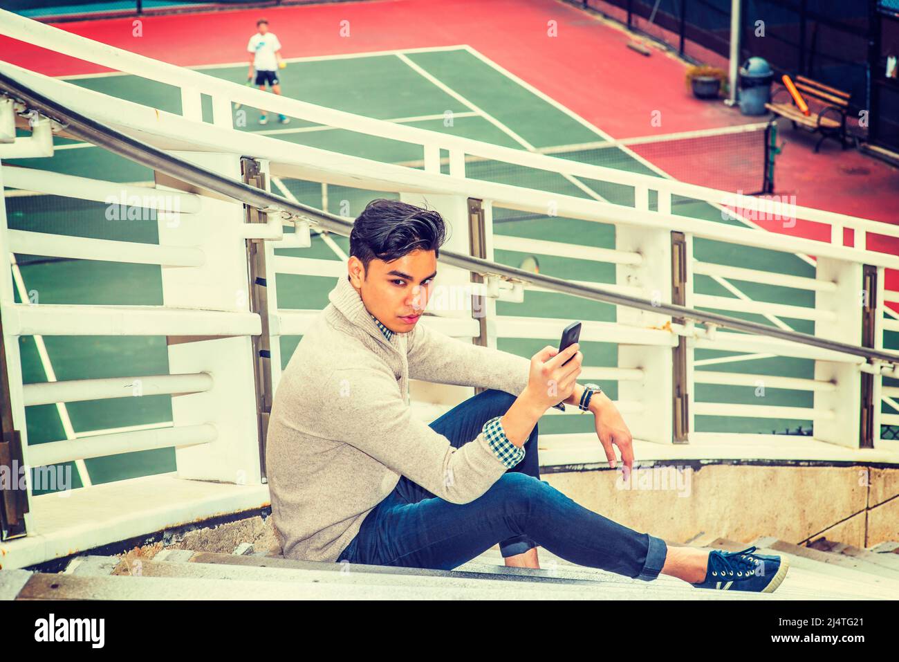 Mann wartet auf dich. Ein junger Mann sitzt auf einer Treppe am Zaun von zehn Mann und kleidet sich in einen Strickpullover mit Schalkragen, Strickjacke, Jeans und über die Manschette Stockfoto